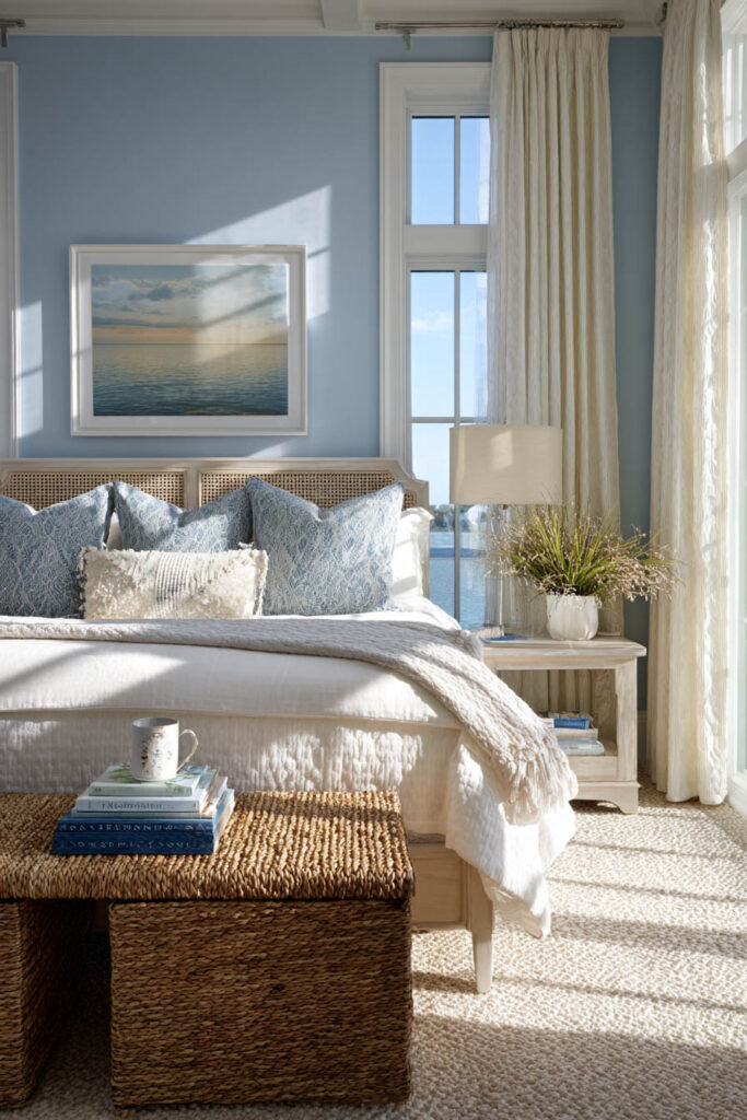 Coastal bedroom with blue walls, white bedding, rattan accents, and ocean artwork in natural sunlight.