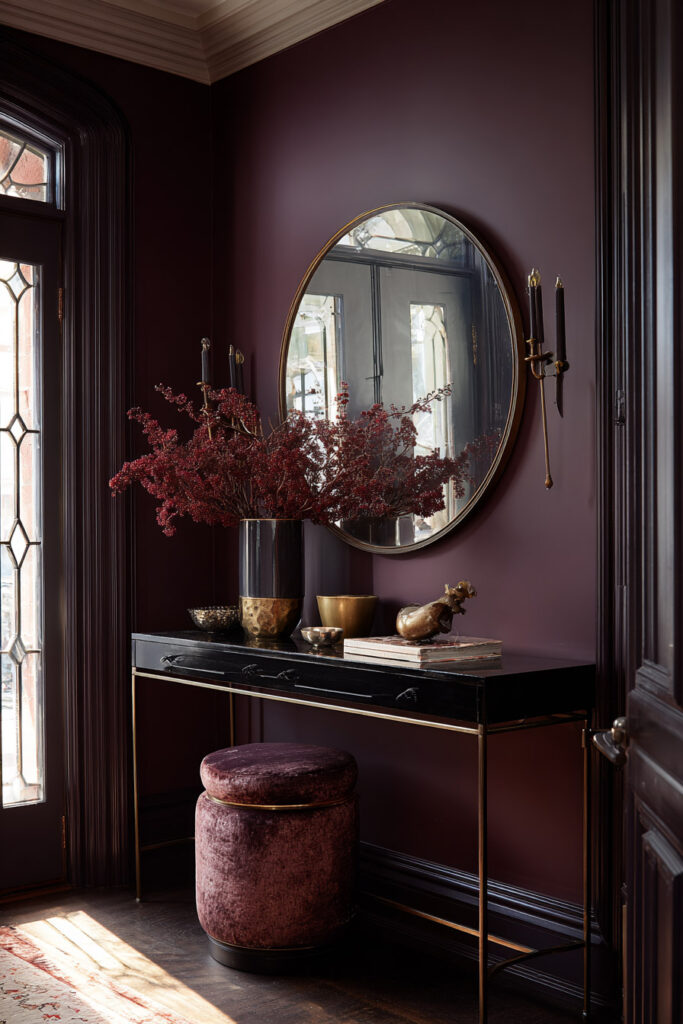Moody plum entryway with black console table, gold decor, round mirror, and velvet stool styled with rich red florals in soft natural light