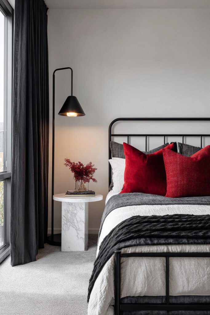 Modern black and white bedroom with red accent pillows, black metal bed frame, marble side table, and moody lighting.