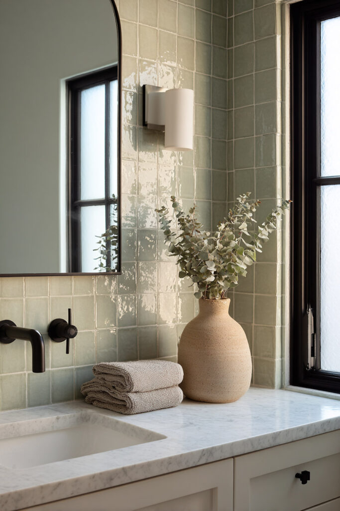 Sage green bathroom with glossy zellige tile backsplash, marble countertop, matte black faucet, and ceramic vase with eucalyptus.