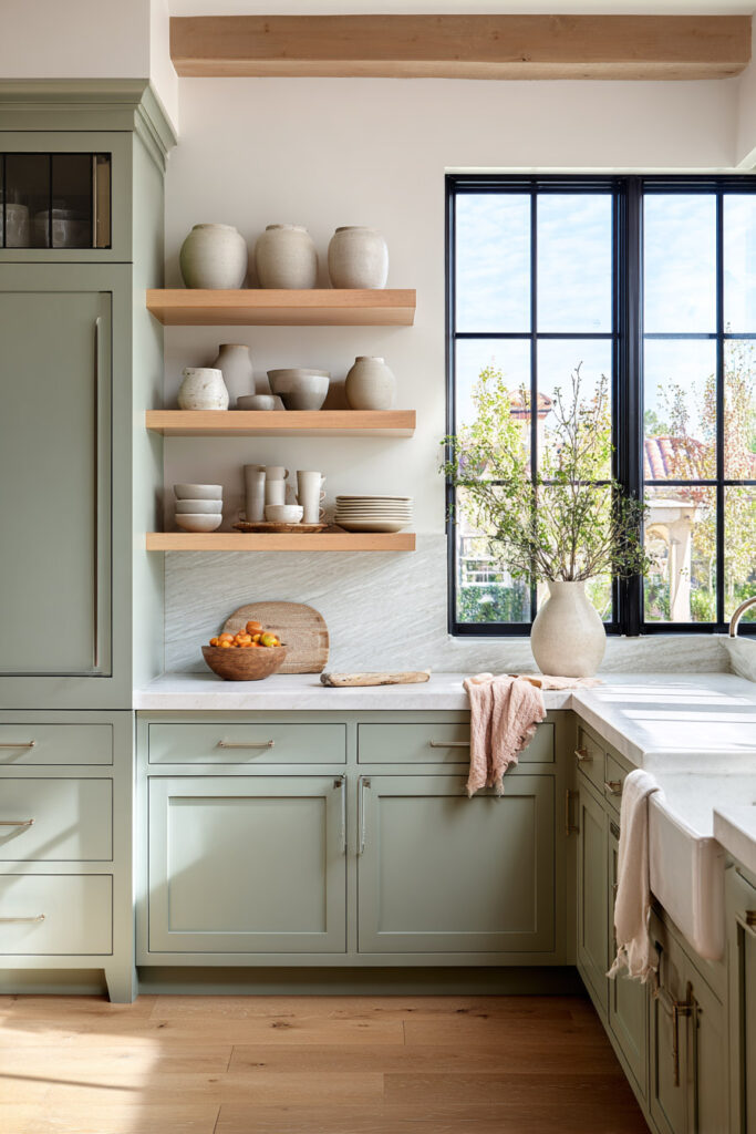 Sage green kitchen with open wood shelves, neutral ceramics, farmhouse sink, and natural light in a modern rustic style.