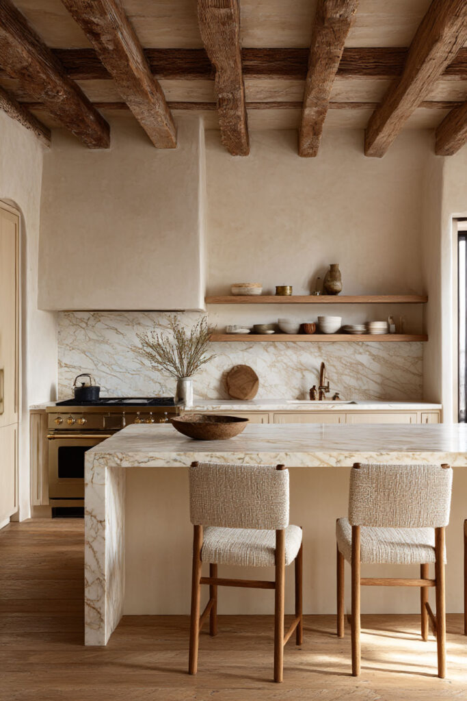 Warm neutral kitchen with plaster walls, rustic wood ceiling beams, marble waterfall island, open shelving, and textured counter stools.