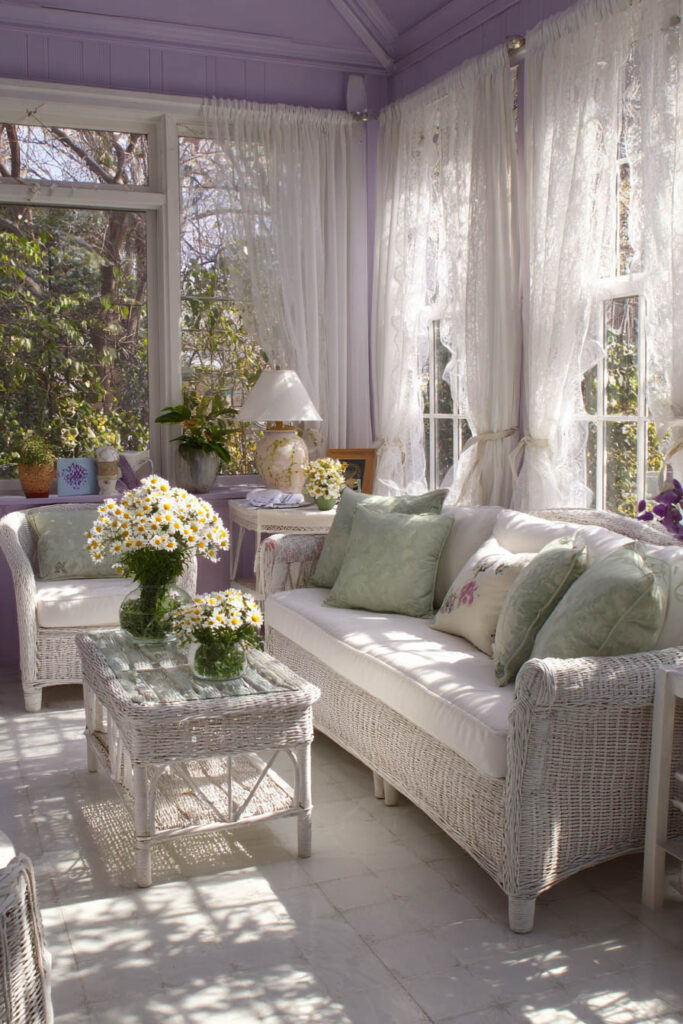 Lavender sunroom with white wicker furniture, green pillows, and vases of daisies in natural light.