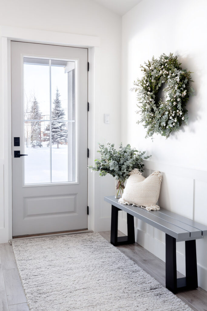 Bright winter entryway with a green wreath, eucalyptus bouquet, and neutral bench decor against a snowy outdoor view.