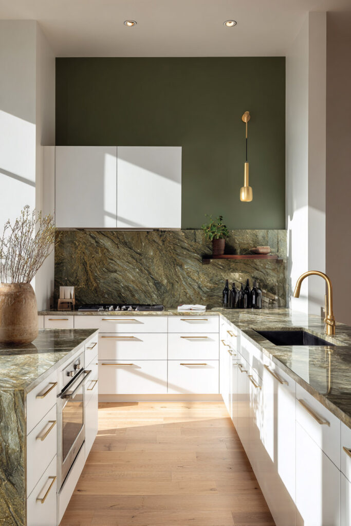 Modern kitchen with olive green stone countertops and backsplash, white cabinetry, brass hardware, and natural sunlight creating a warm, elegant contrast.