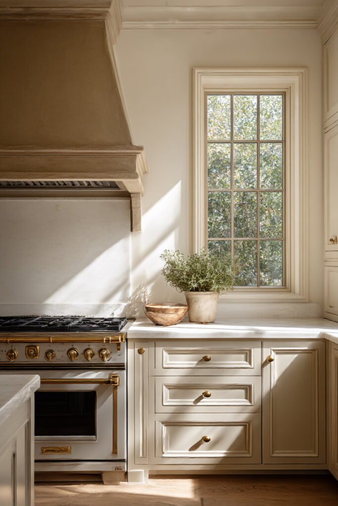 Warm neutral kitchen with brass-accented range, creamy cabinets, marble countertops, plaster hood, sunlit window, and rustic decor.