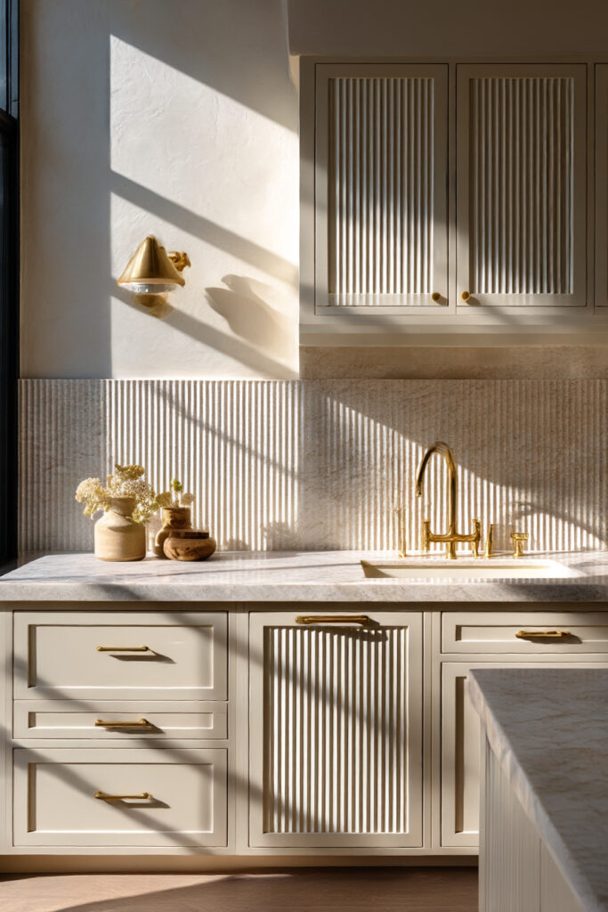 Kitchen with textured stone or clay tile backsplash, vertical ribbed tiles, cream cabinets, fluted cabinet fronts, and brass fixtures.