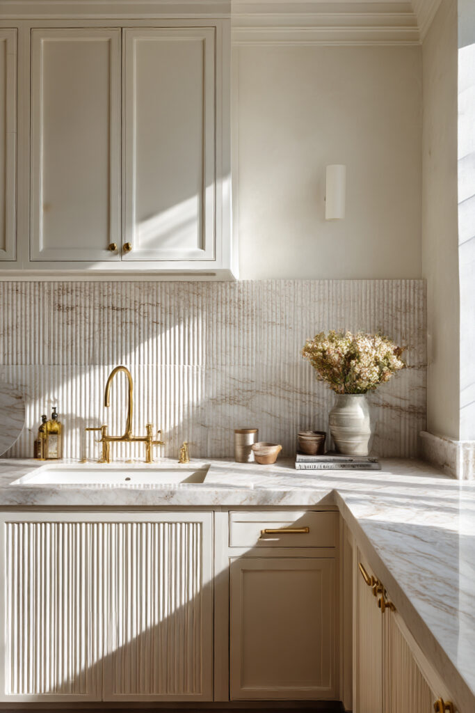 Textured stone kitchen backsplash with vertical fluting, marble countertops, cream cabinets, and brass hardware