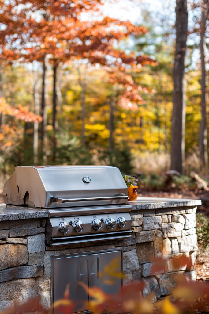 Outdoor grill station with built-in stainless steel grill, natural stone base, and granite countertop set in a wooded backyard.