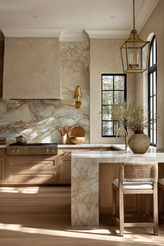 Kitchen with veined stone backsplash, plaster range hood, warm wood cabinets, brass lighting, and natural stone island.