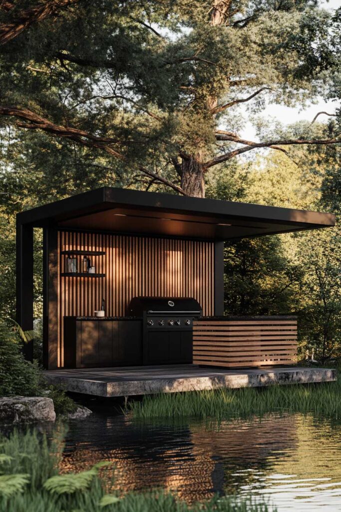 Modern outdoor grill station with black cabinetry, wood slat walls, flat roof, and waterfront forest setting