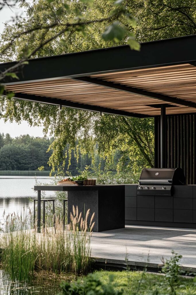 Modern outdoor grill station with black cabinets, built-in gas grill, pergola roof, and lakeside backyard setting.