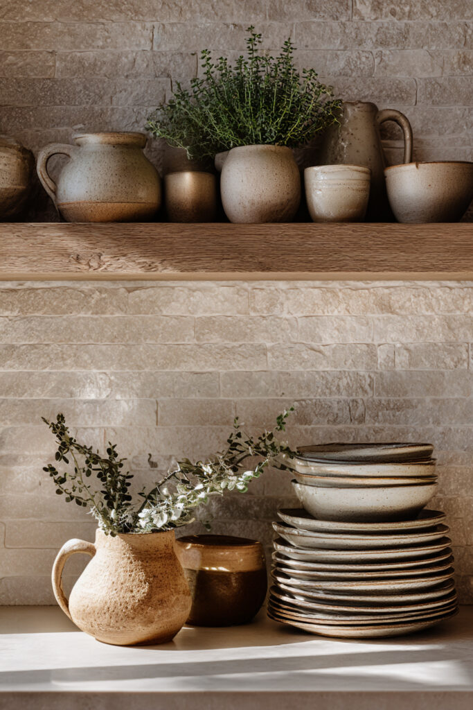 Kitchen with stone or clay tile backsplash, open wood shelves, handmade ceramic dishes, rustic pottery, and neutral styling.