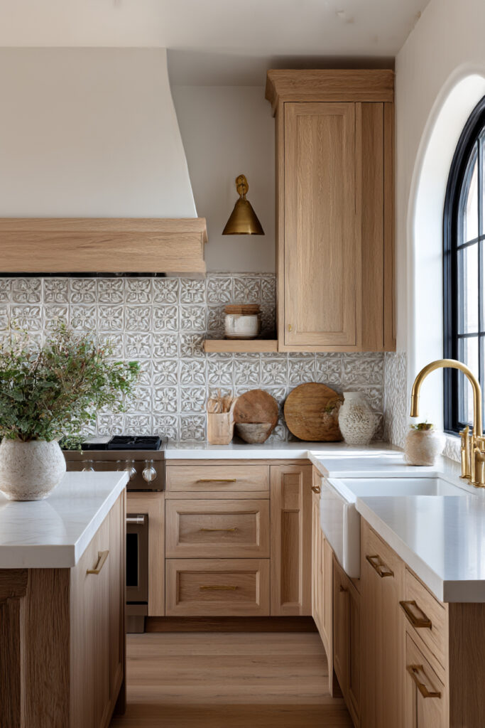 Kitchen with textured stone tile backsplash, warm brass wall sconce, light wood cabinetry, and farmhouse sink in an earthy neutral palette.