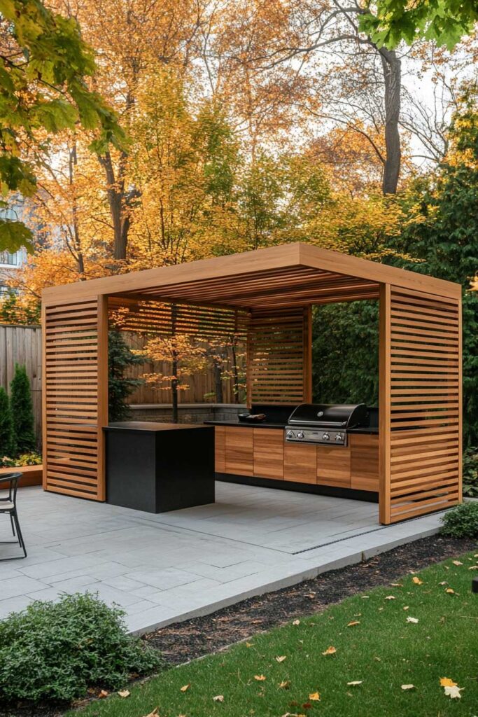 Modern outdoor grill station with wood slat pergola, built-in grill, black countertop, and wood cabinetry in a fall backyard.