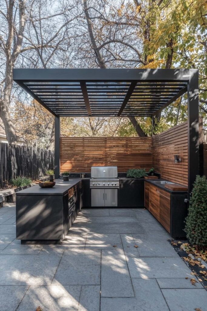 Modern outdoor grill station with black pergola, wood privacy panels, built-in stainless steel grill, and stone patio flooring.