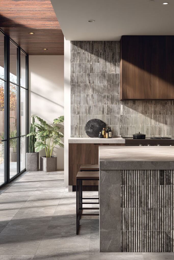 Kitchen with ribbed backsplash, dark wood island, large potted plants, and sunlight highlighting textured surfaces
