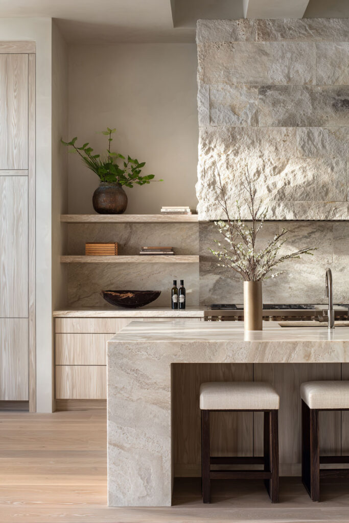 Modern neutral kitchen with textured stone backsplash, thick stone island, and natural wood cabinetry highlighting dimensional surface trends and organic kitchen materials.