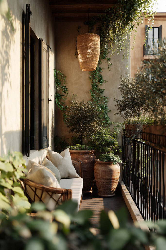 Small Mediterranean balcony with layered greenery, olive trees, climbing vines, terracotta planters, woven pendant light, and private outdoor seating area.