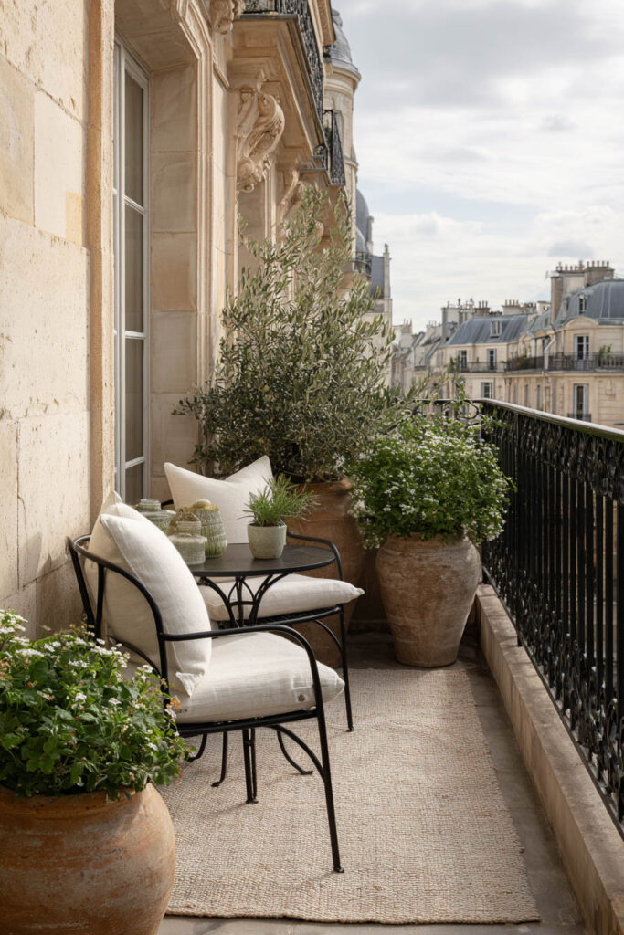 Parisian balcony with neutral outdoor furniture, olive tree, terracotta planters, cream cushions, and soft natural textures overlooking classic French buildings.