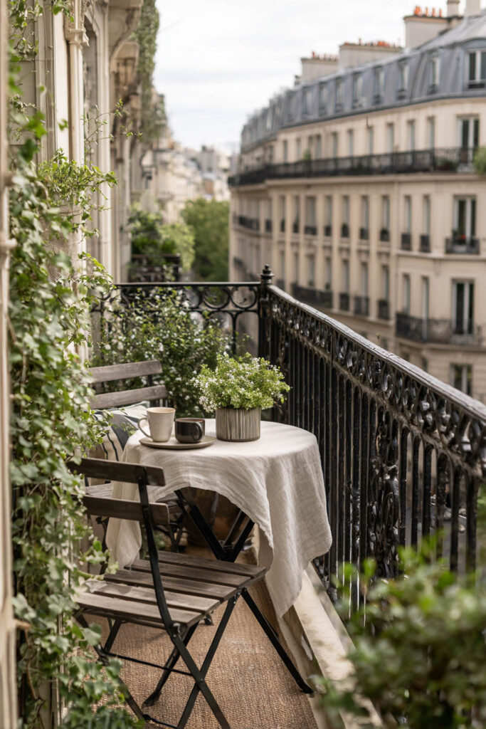 Small Parisian balcony with a classic wooden bistro set, linen-covered table, climbing greenery, wrought iron railing, and elegant city views.