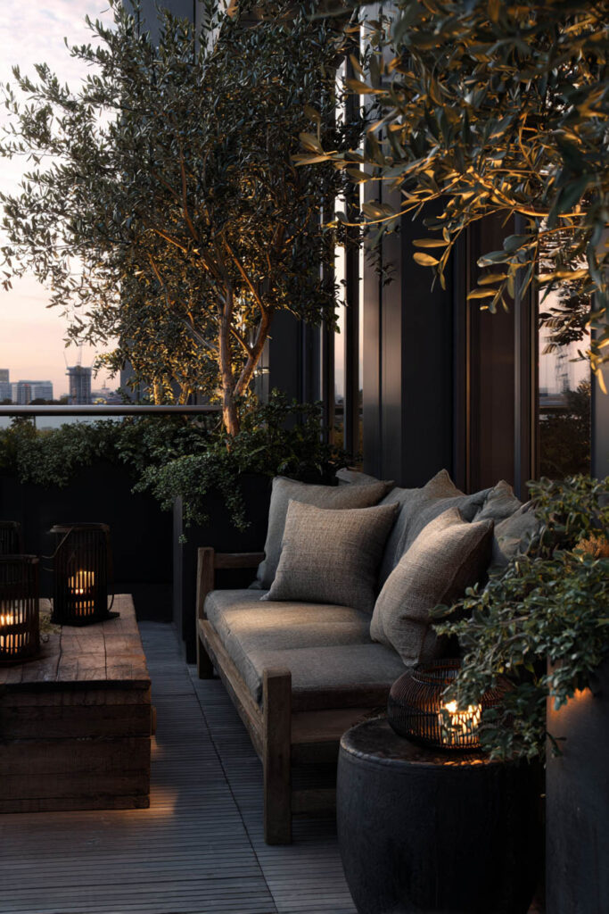 Dark apartment balcony with lush greenery, cozy outdoor sofa, warm lantern lighting, and layered plants creating a private retreat.